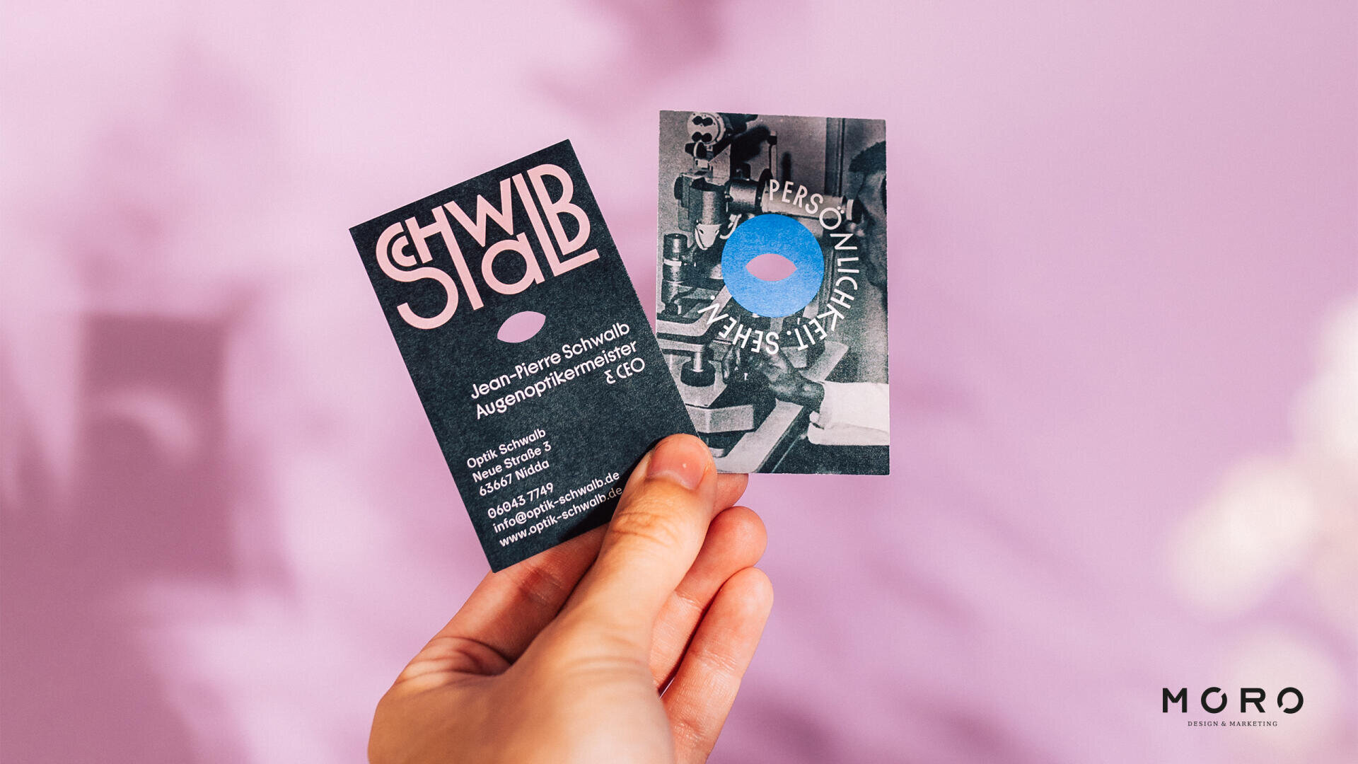 Eine Hand hält zwei Visitenkarten vor einem zartvioletten Hintergrund. Die linke Karte enthält fettgedruckten, stilisierten Text und Kontaktinformationen; die rechte Karte zeigt abstrakte Kunst mit einem blauen Kreis und teilweisem Text. Das MGRO-Logo befindet sich in der unteren rechten Ecke.