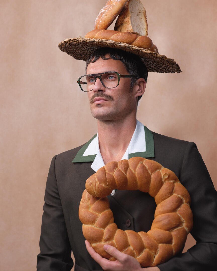 A man with glasses and a mustache wears a suit and a woven hat topped with bread, while holding a large braided bread wreath against a beige background.