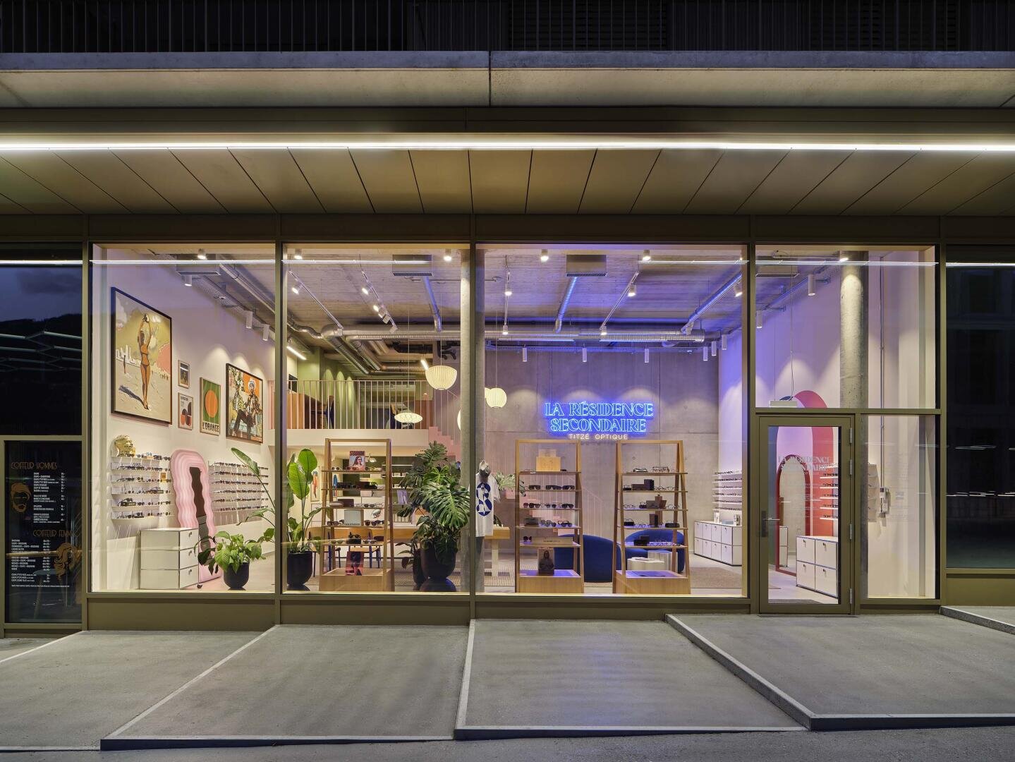 Ein modernes Brillengeschäft mit großen Glasfenstern, hell erleuchtetem Interieur, Regalen mit Brillen, Pflanzen, bunten Kunstwerken und einem blauen Neonschild mit der Aufschrift LA RÉSIDENCE SPÉCIALITÉS.