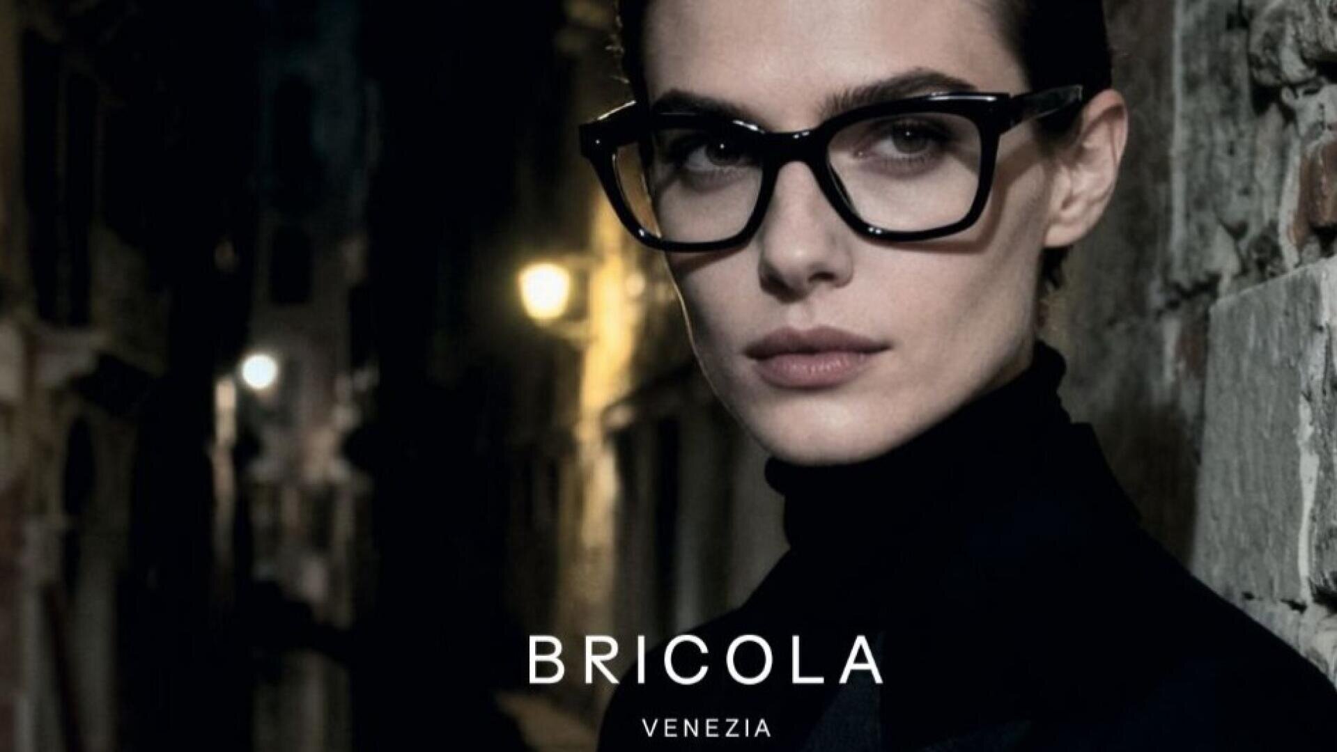 A person with short dark hair and large black glasses stands against a dimly lit alleyway at night. The word BRICOLA with VENEZIA below it appears at the bottom of the image.