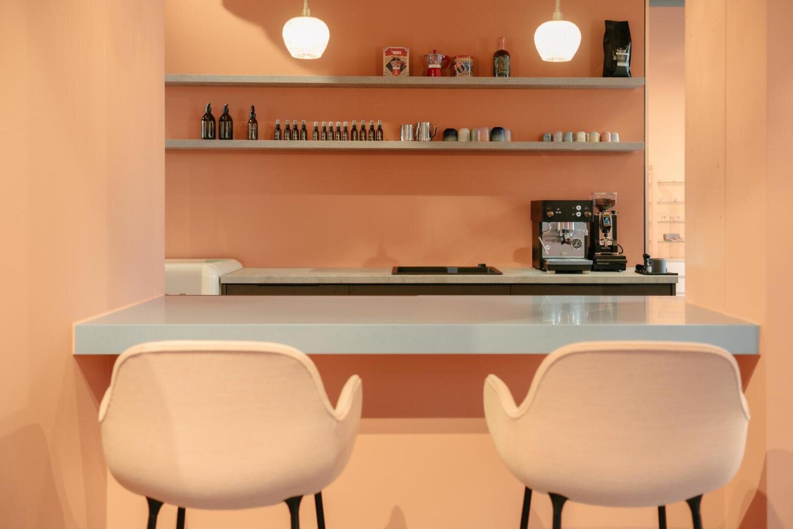Minimalist peach-colored café interior with two light pink chairs at a gray counter, shelves displaying coffee items, jars, and bottles, with a coffee machine and cups in the background.