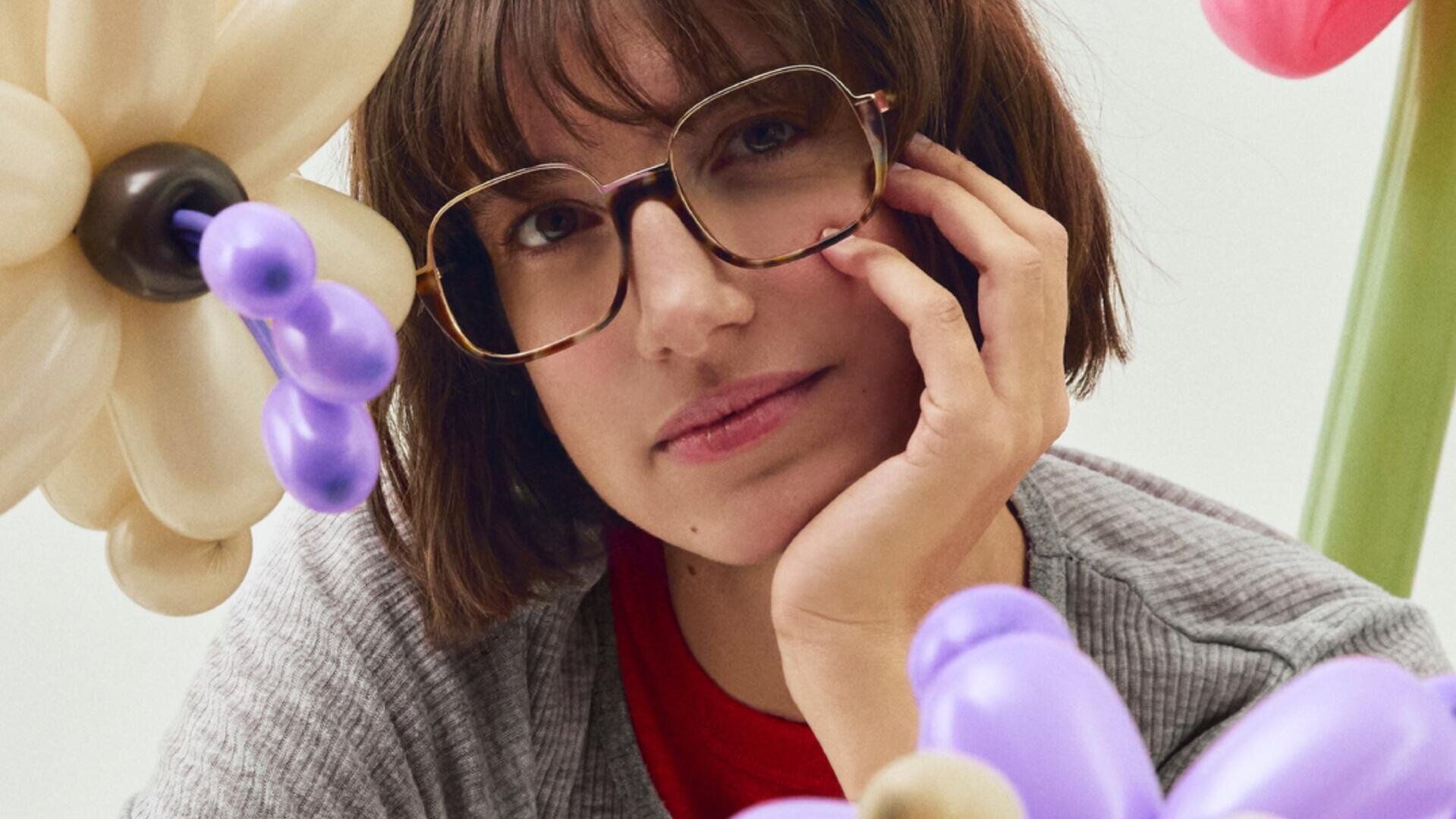 A woman with brown hair and glasses rests her face on her hand, surrounded by colorful balloon flowers in purple, cream and green. She wears a gray sweater over a red shirt and looks thoughtfully into the camera.