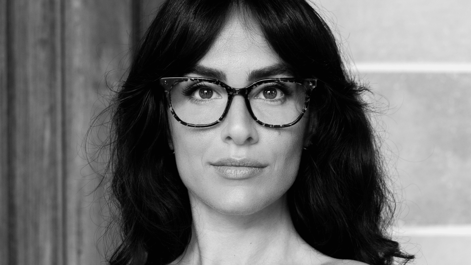 Schwarz-Weiß-Porträt einer Frau mit dunklem gewelltem Haar, die eine gemusterte Brille trägt und mit neutralem Blick direkt in die Kamera schaut. Der Hintergrund ist unscharf.