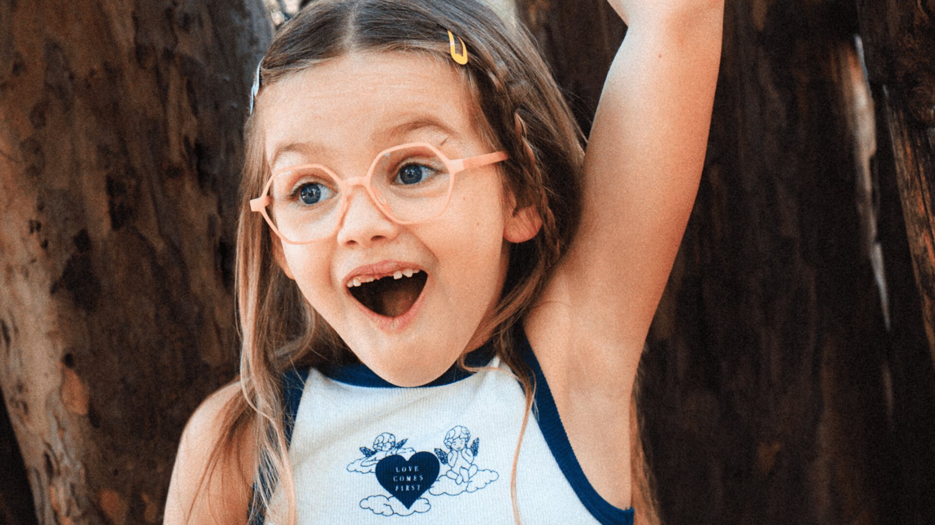 Ein junges Mädchen mit langen Haaren, einer hellrosa Brille und einem weißen Tanktop steht aufgeregt lächelnd mit offenem Mund und erhobenem Arm vor einem Baum.