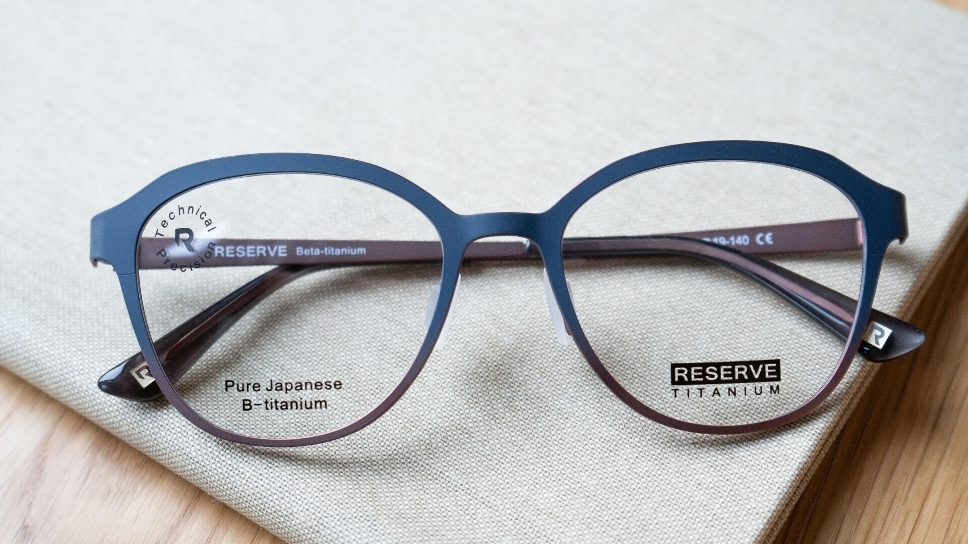 Eine runde Brille mit dunklem Rahmen und den Worten „Pure Japanese B-Titanium“ und „RESERVE TITANIUM“ auf den Gläsern, die auf einer hellbeigen strukturierten Oberfläche ruhen.