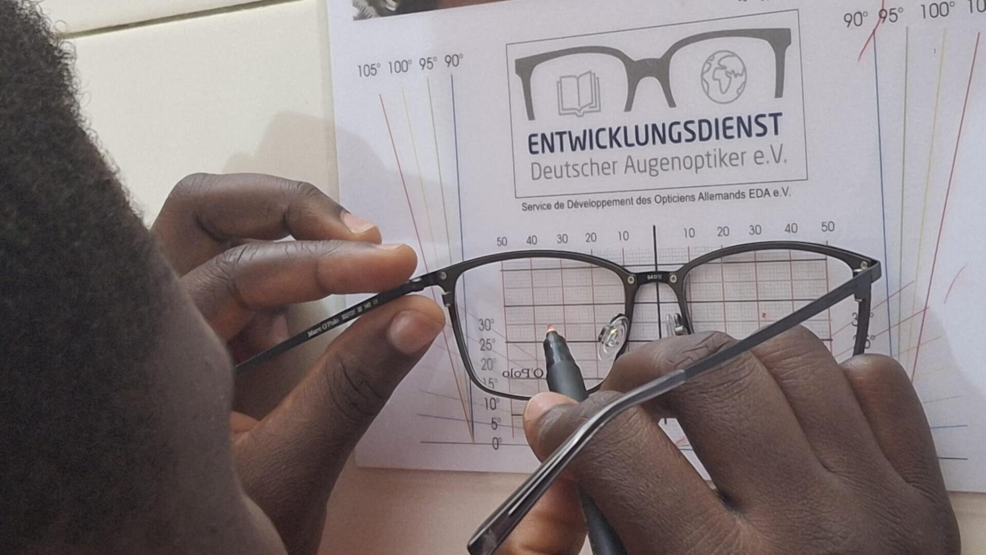 A person measures spectacle frames with a ruler and aligns them according to a table labeled "ENTWICKLUNGSDIENST Deutscher Augenoptiker e.V." and angle markings.
