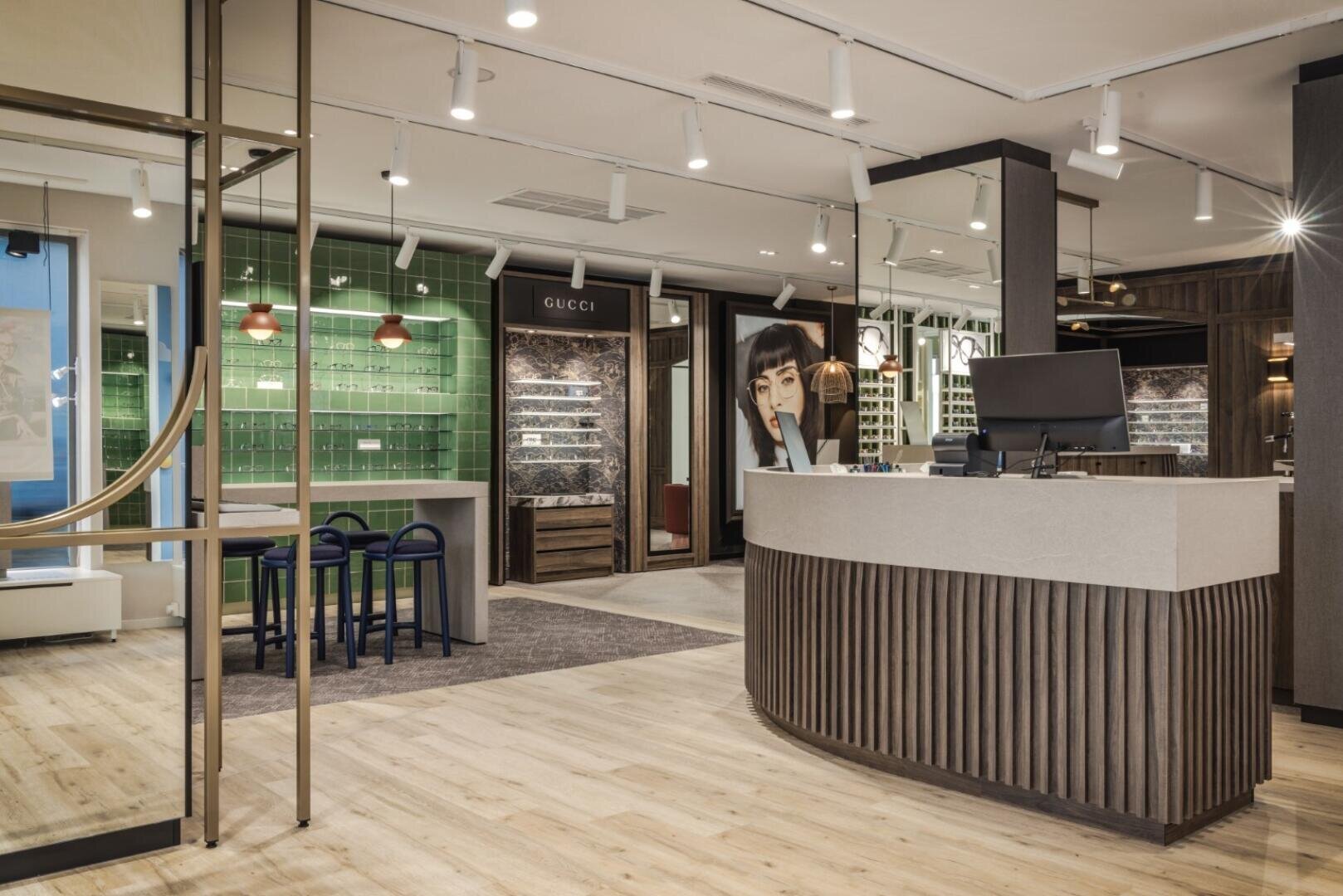 Modernes Optikergeschäft mit stilvoller Beleuchtung, Holzböden, grün gefliesten Wänden, Markenregalen, einem geschwungenen Empfangstresen, einem Sitzbereich und verschiedenen Brillenauslagen.