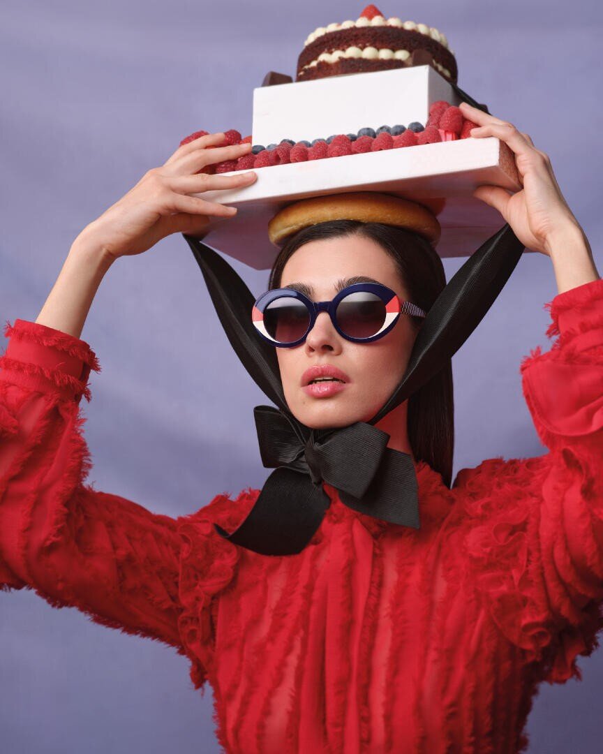 A woman in a red, structured dress and round sunglasses balances a cake and pastry on her head, secured with a large black ribbon tied under her chin, against a light purple background.