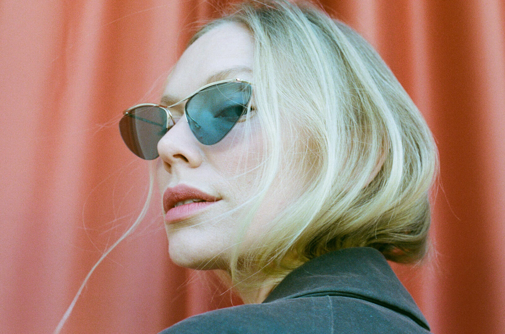 A person with short blond hair, wearing sunglasses with cat eyes and a dark jacket, looks over her shoulder in front of a coral-pink curtain.