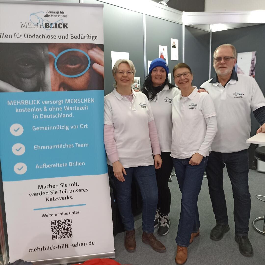 Four smiling people in white multi-vision shirts stand next to a blue and white information banner about free eye care services for the homeless and needy in Germany. They appear to be at an indoor event or trade fair.