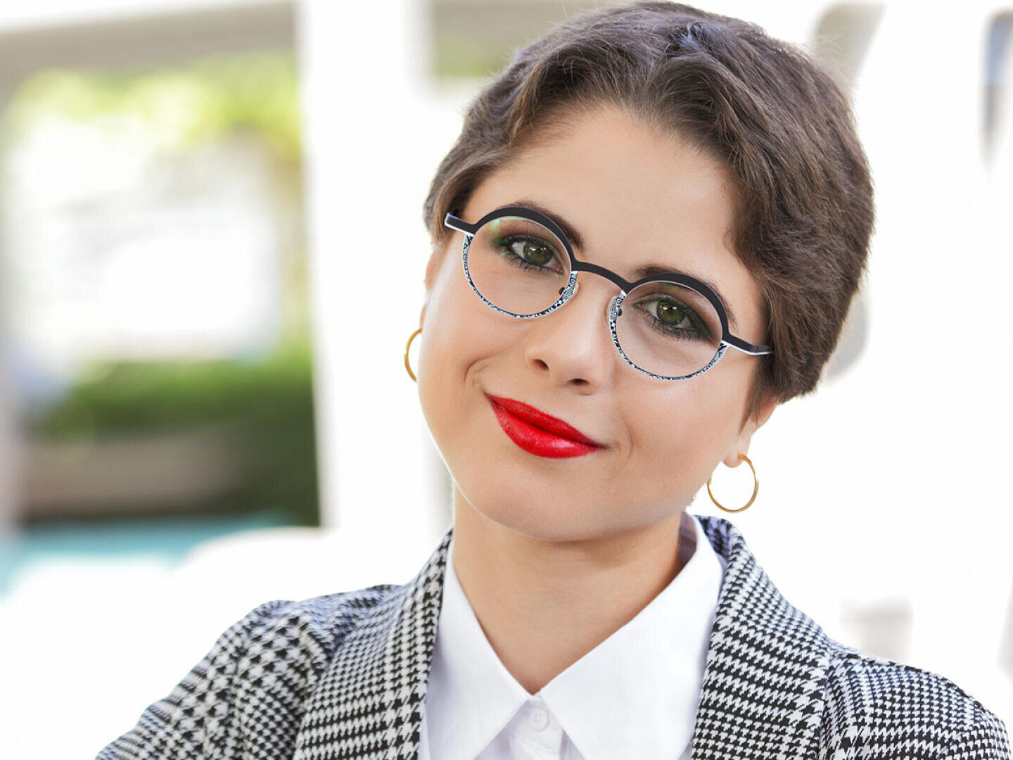 A person with short brown hair, round glasses, red lipstick, and gold hoop earrings smiles slightly while wearing a black-and-white checkered blazer and white shirt, with a blurred outdoor background.