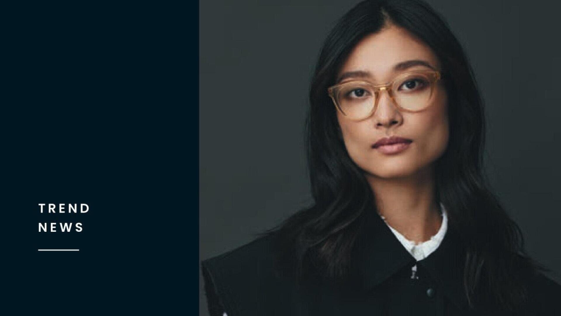 A woman with long dark hair and glasses looks at the camera with a neutral expression. She wears a black jacket over a white top. The left side of the image features a dark panel with the text TREND NEWS.