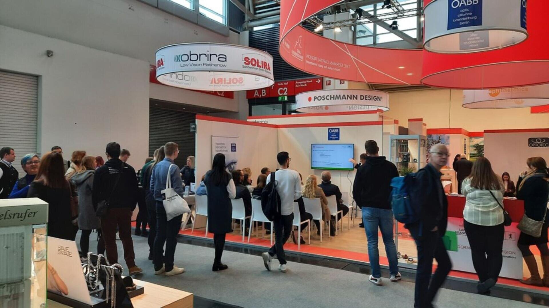 A group of people gathers around a presentation area at a busy trade show or exhibition, with displays, banners, and seating for attendees visible in a modern indoor setting.