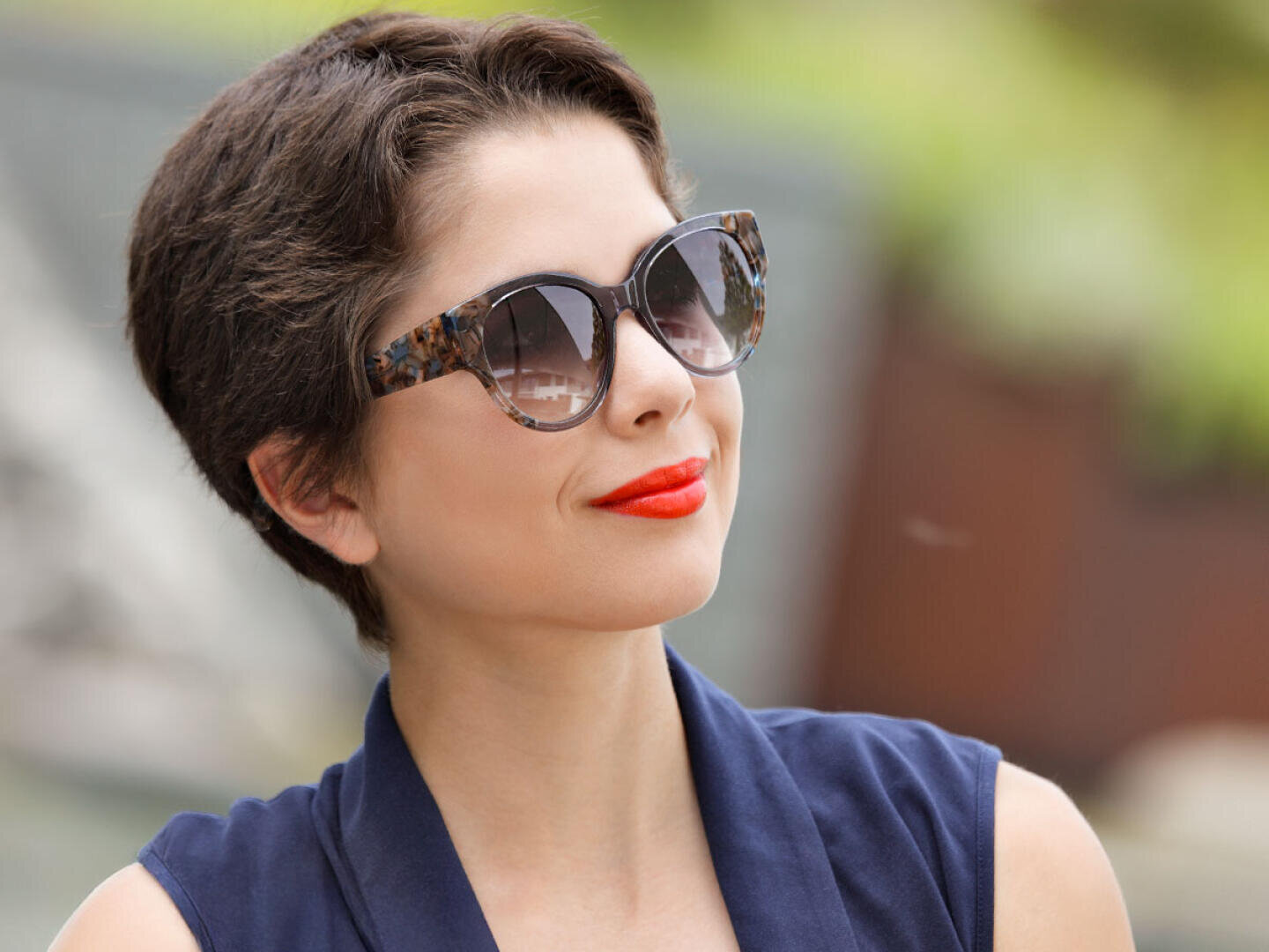 Eine Frau mit kurzen braunen Haaren trägt eine Schildpatt-Sonnenbrille und leuchtend roten Lippenstift. Sie trägt ein ärmelloses marineblaues Oberteil und lächelt im Freien vor einem unscharfen grünen Hintergrund.