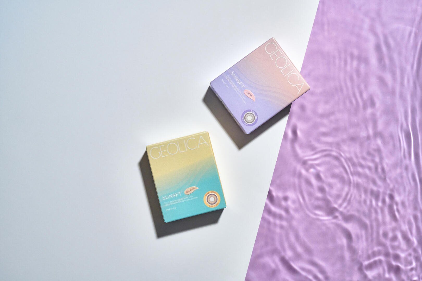 Two pastel-colored GEOLICA Sunset contact lens containers are arranged on a surface, with one on a plain white surface and the other near a purple rippled section of water, creating a soft and modern aesthetic.