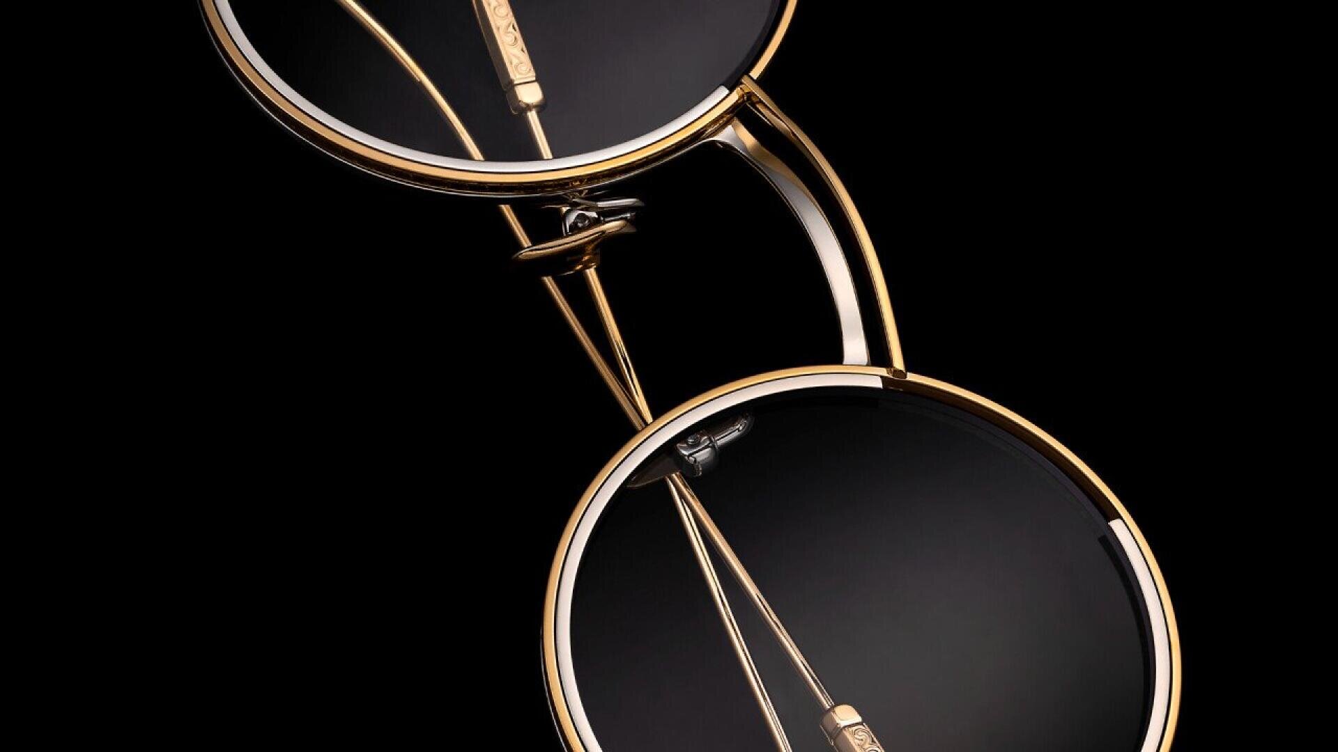Close-up of two pairs of round eyeglasses with gold frames and black lenses, arranged diagonally on a black background. The glasses’ arms and intricate frame details are clearly visible.