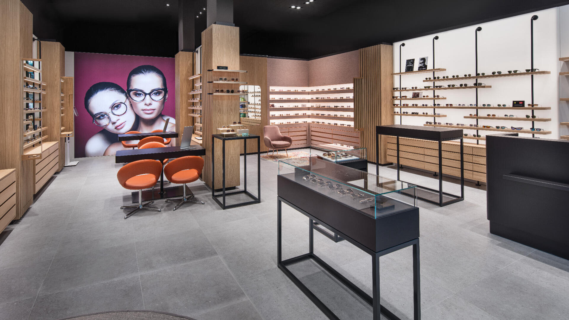 Modernes Brillengeschäft mit Holzregalen, in denen verschiedene Brillengestelle ausgestellt sind, orangefarbenen Stühlen, einer Glasvitrine und einem großen Wandfoto von zwei Frauen mit Brillen. Der Raum ist hell und übersichtlich.