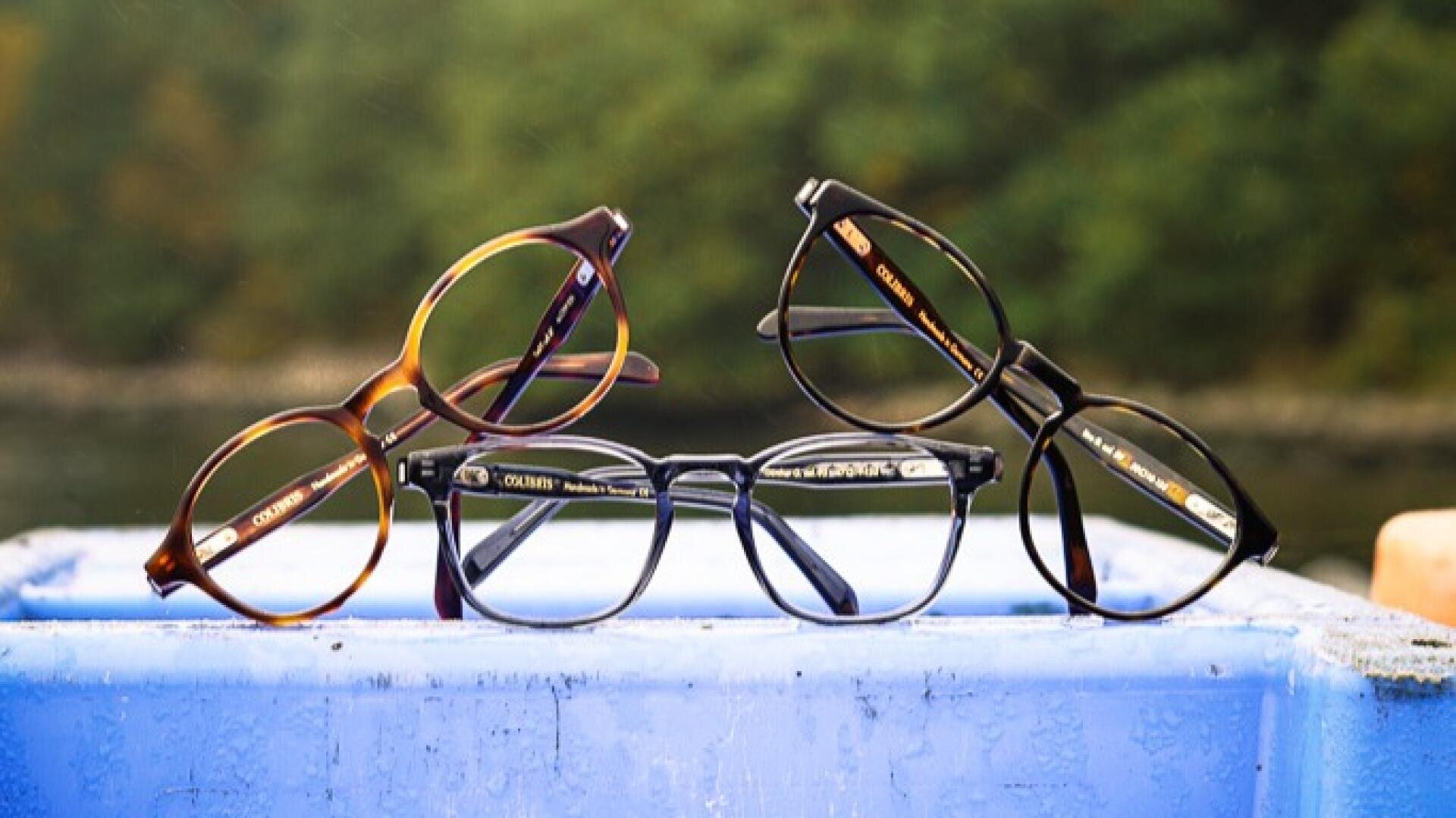 Drei Brillen mit verschiedenfarbigen Rahmen (braun, schwarz und klar) werden auf einer blauen Kunststoffoberfläche mit einem unscharfen grünen Hintergrund im Freien ausgestellt.