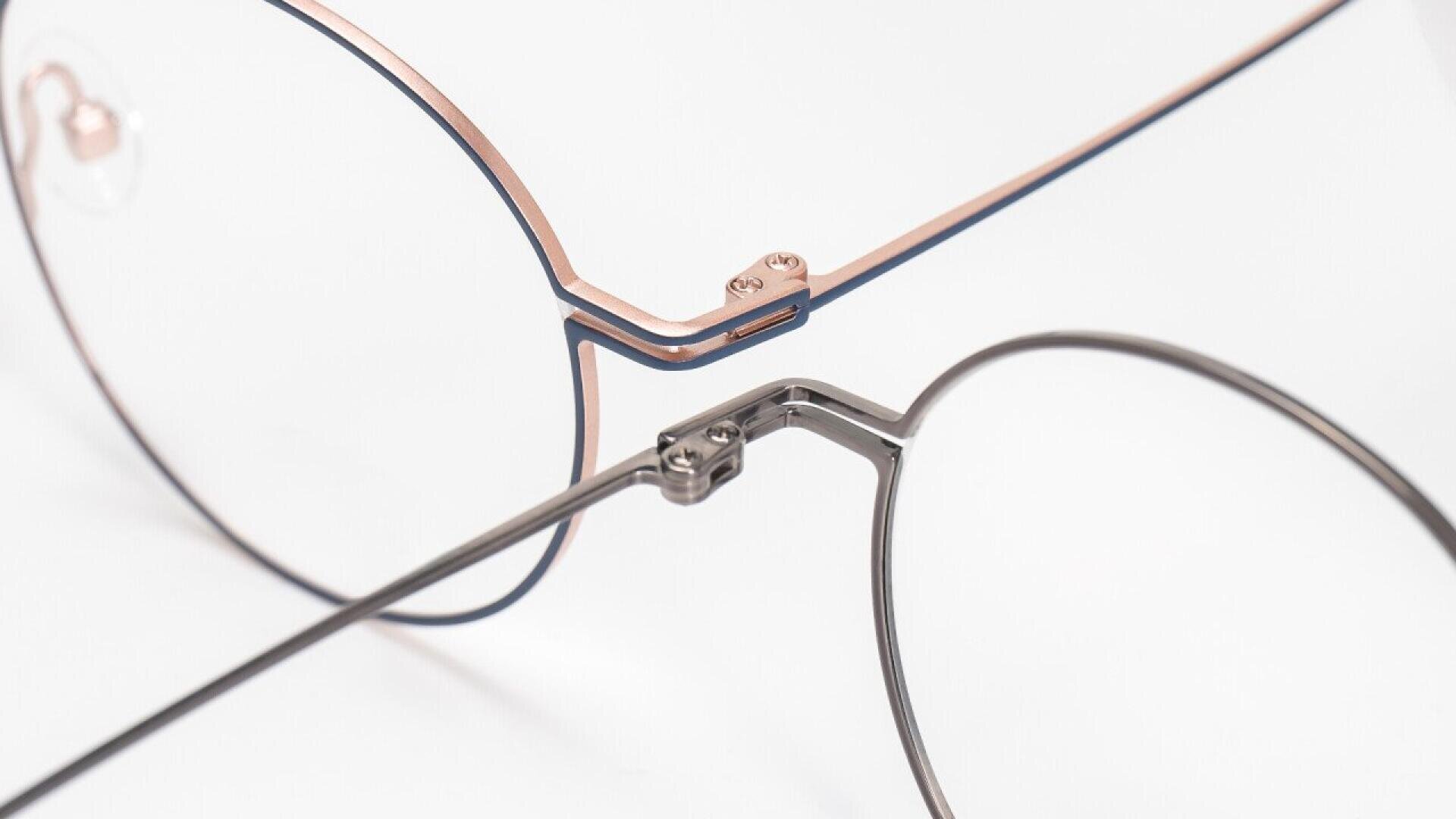 Close-up of the bridge and frames of a pair of eyeglasses with thin, metallic rims and clear lenses on a white background. The frame has a modern, minimalistic design.