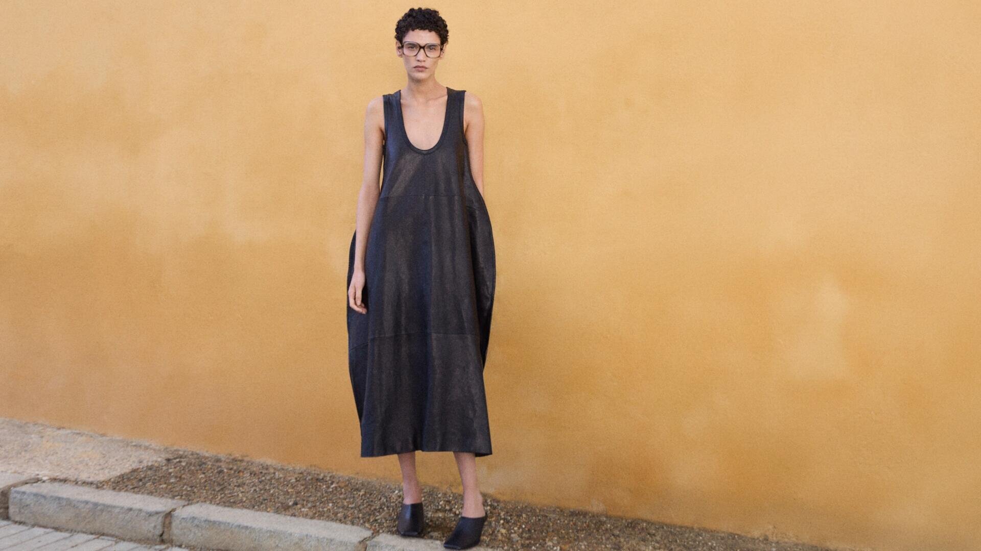 Eine Person mit kurzen lockigen Haaren und Brille steht in einem lockeren, ärmellosen, dunklen Kleid und schwarzen Schuhen auf einem Bürgersteig mit Kies und Pflastersteinen vor einer schlichten beigen Wand.