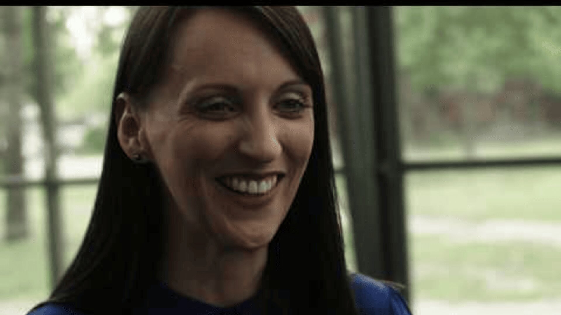 Eine Frau mit langem, dunklem Haar sitzt lächelt drinnen an großen Fenstern, im Hintergrund sind draußen grüne Bäume und Gras zu sehen.