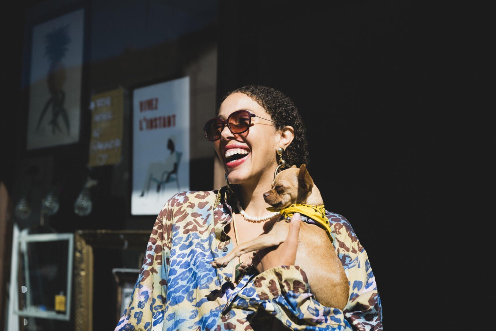 Eine lächelnde Person mit Sonnenbrille und gemustertem Hemd hält einen kleinen braunen Hund mit einem gelben Geschirr, der draußen im Sonnenlicht steht, während im Hintergrund dunkle Schaufenster und Kunstplakate zu sehen sind.