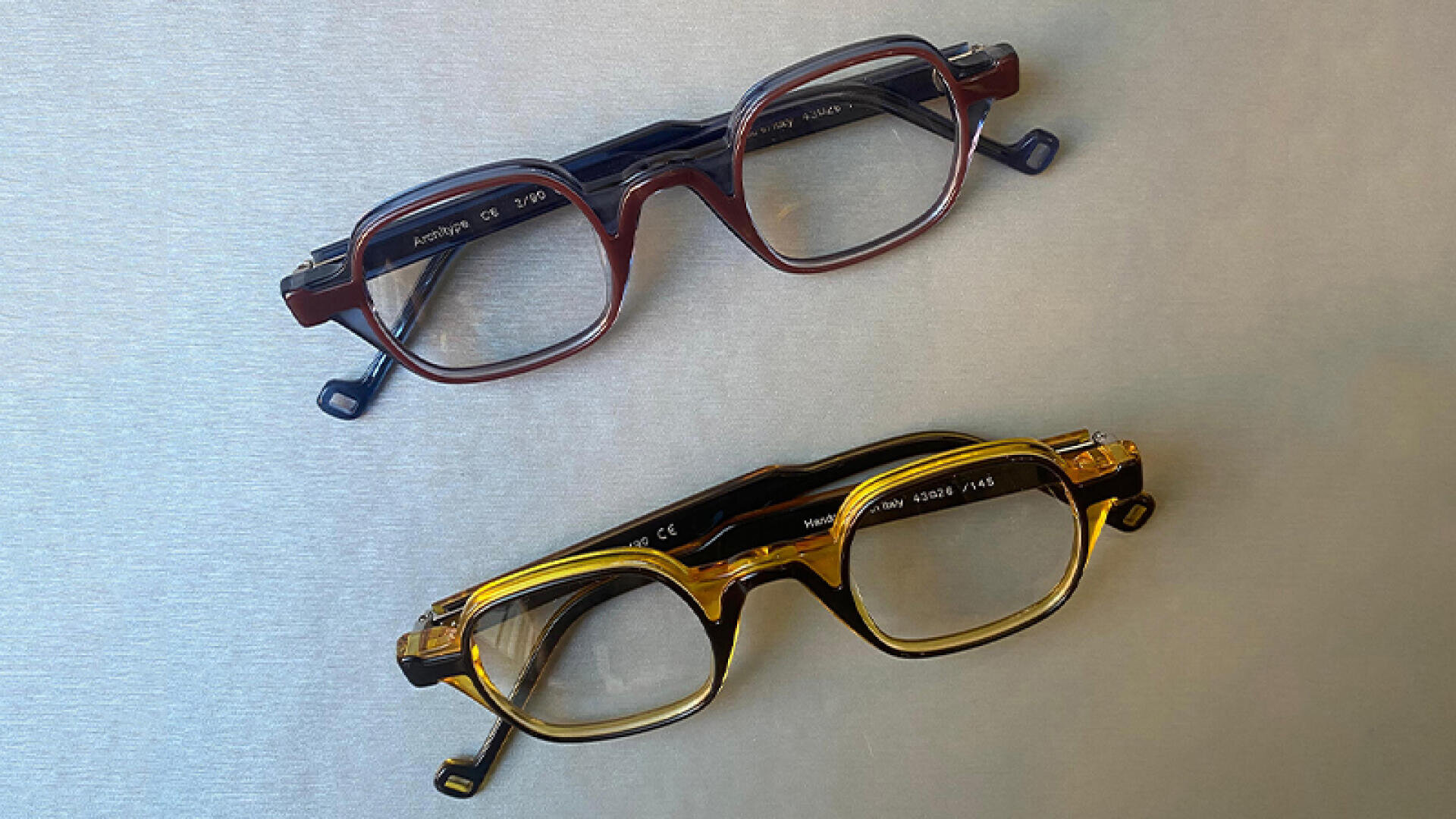 Two pairs of glasses with thick frames - one brown and one yellow - stand on a light gray surface, diagonally and parallel to each other.