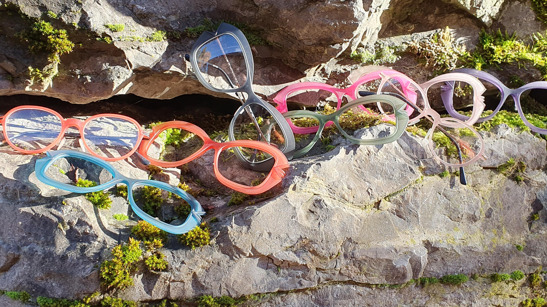 Mehrere bunte Brillengestelle, darunter in Rot, Grün, Rosa und Grau, sind im Freien auf einer sonnenbeschienenen, moosbedeckten Felsoberfläche ausgestellt.