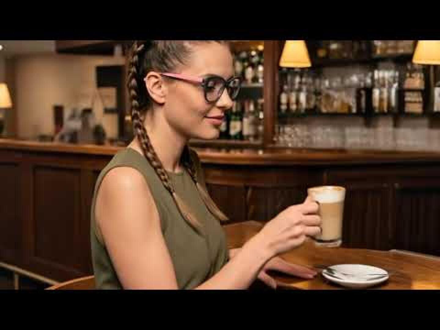 Eine Frau mit geflochtenen Haaren und Brille sitzt an einer hölzernen Bar, in der Hand eine Tasse Kaffee oder Milchkaffee, im Hintergrund Regale mit Flaschen und warmes Licht.