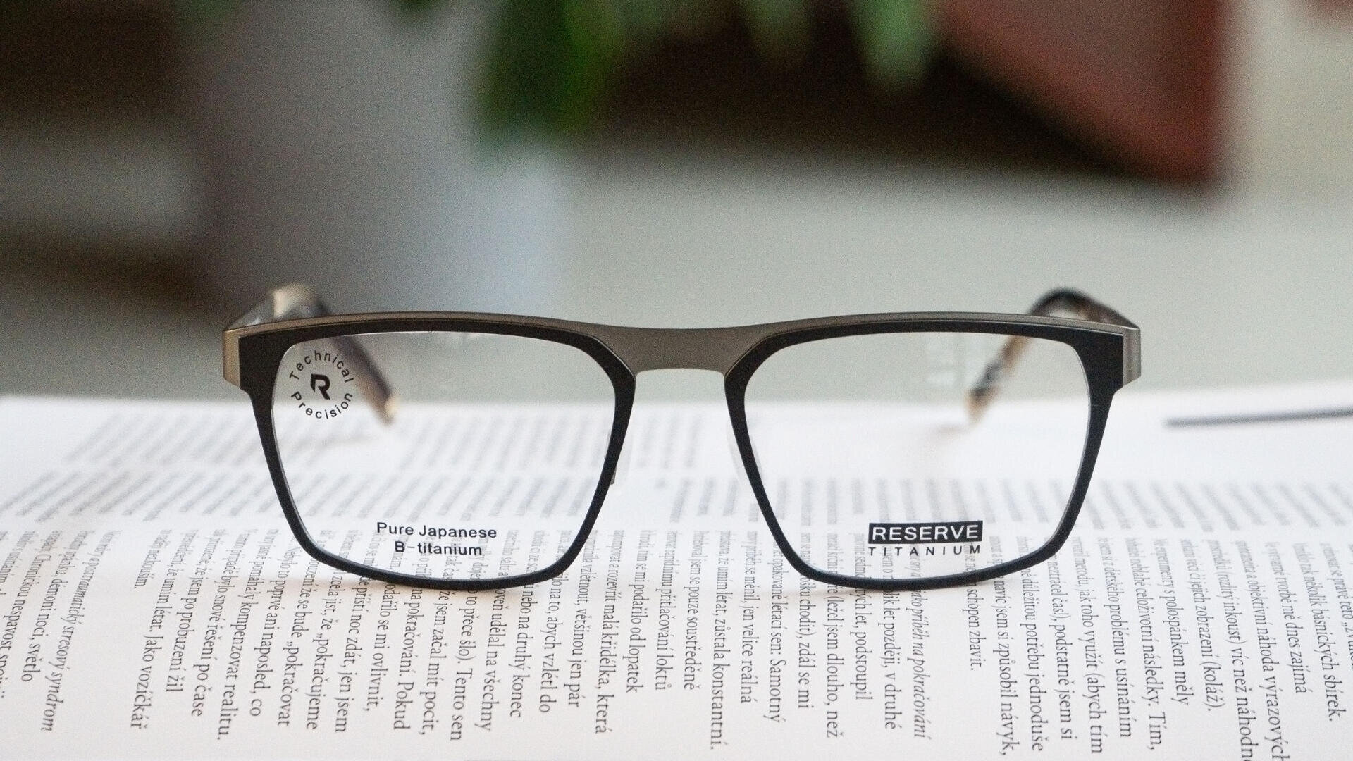 A pair of rectangular eyeglasses with black frames rests on an open book; text through the lenses reads Pure Japanese β-Titanium and RESERVE TITANIUM.