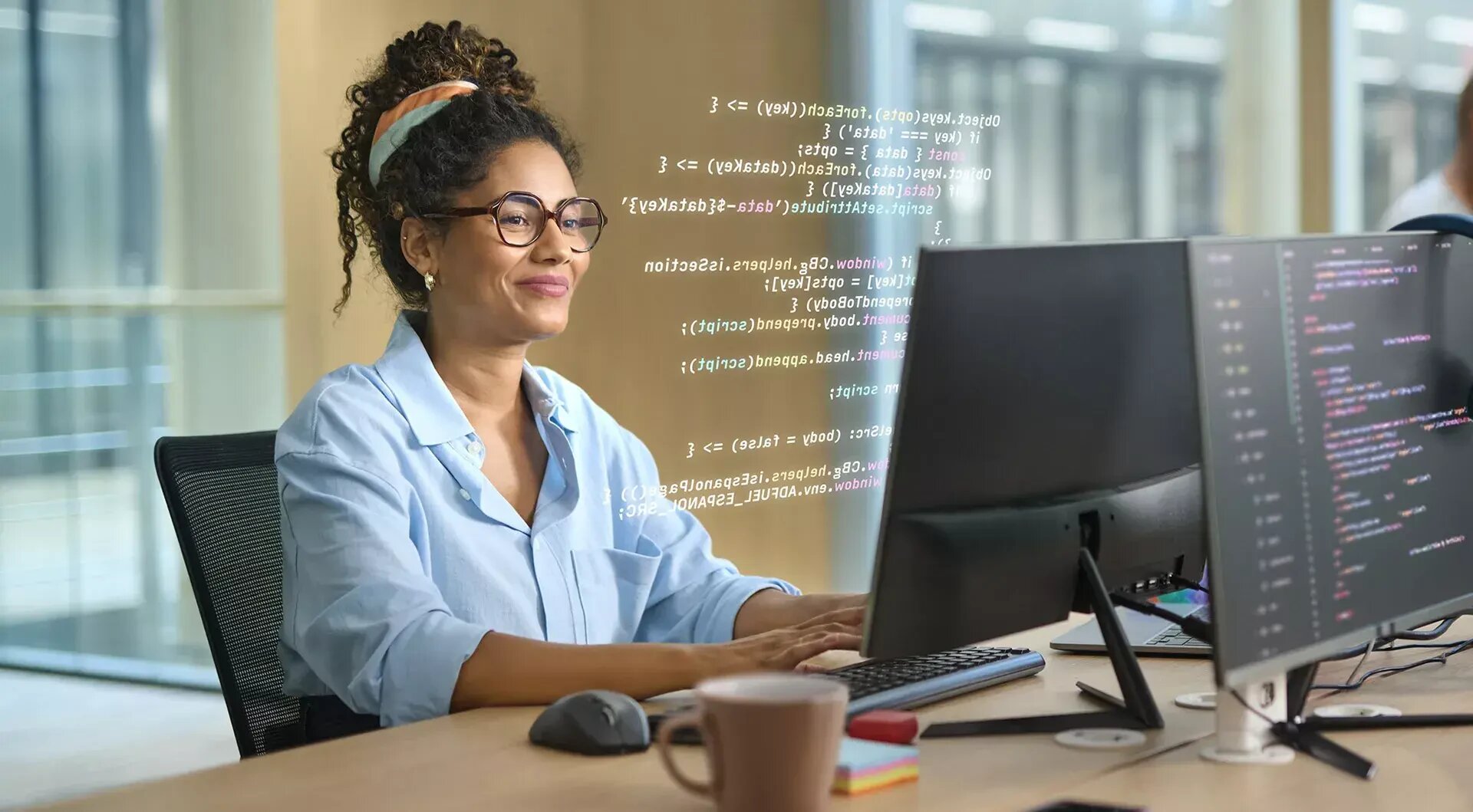 Eine Frau mit Brille und blauem Hemd arbeitet in einem Büro an einem Computer. Auf ihrem Bildschirm ist Code zu sehen, auf dem Schreibtisch steht eine Kaffeetasse. Sie wirkt konzentriert und lächelt leicht.