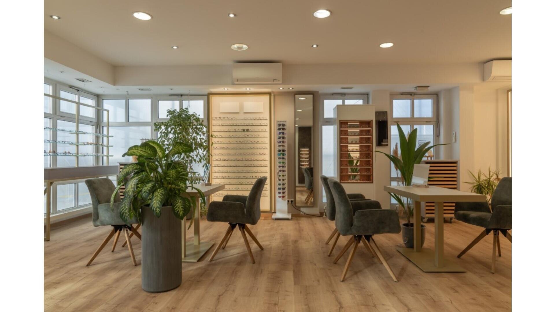 A modern optician’s office with large windows, green indoor plants, display shelves with eyeglass frames, and contemporary seating around sleek tables on a light wood floor.