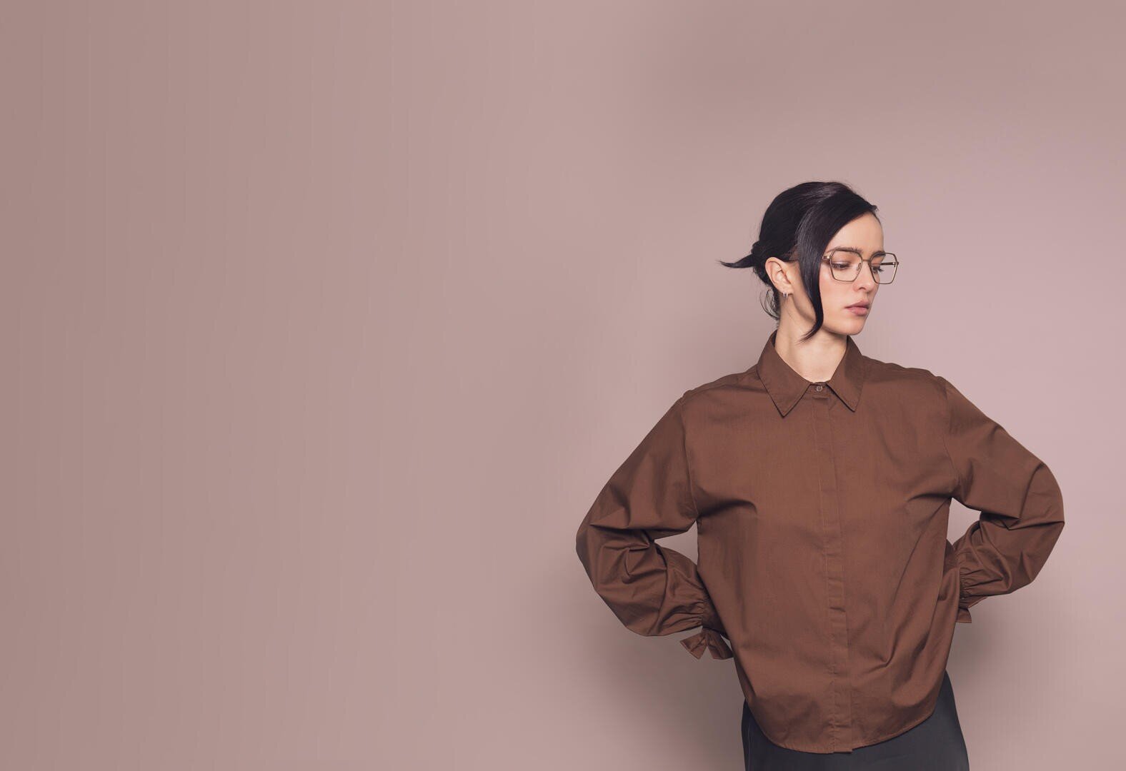 Eine Person mit Brille und dunklem Haar, die ein braunes Hemd trägt, steht vor einem schlichten hellbraunen Hintergrund, die Hände in die Hüften gestemmt und schaut zur Seite.