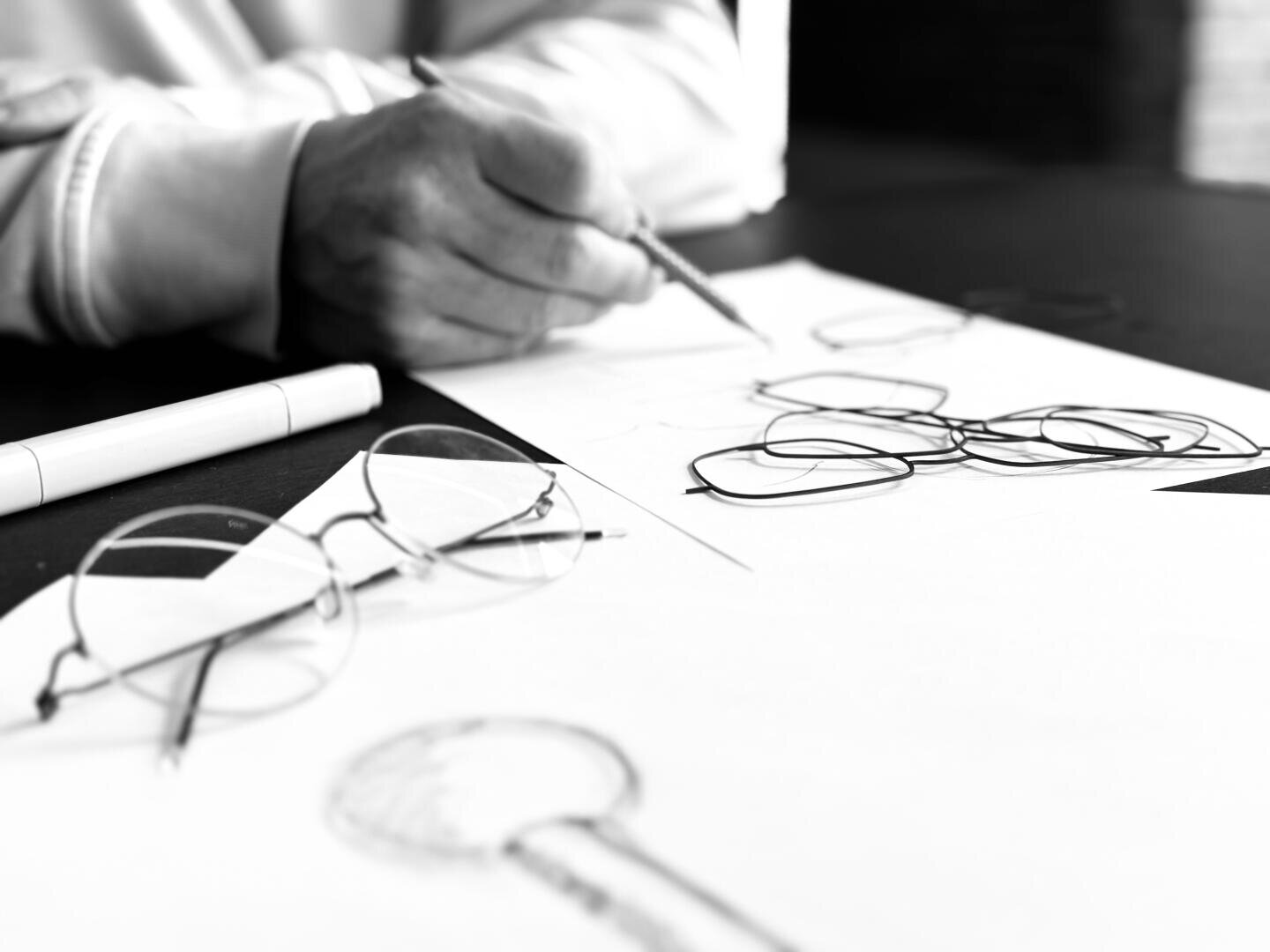 Eine Person skizziert mit einem Bleistift Brillengestelle auf Papier; auf dem Tisch liegen eine Brille und ein Marker, alles in schwarz-weiß.