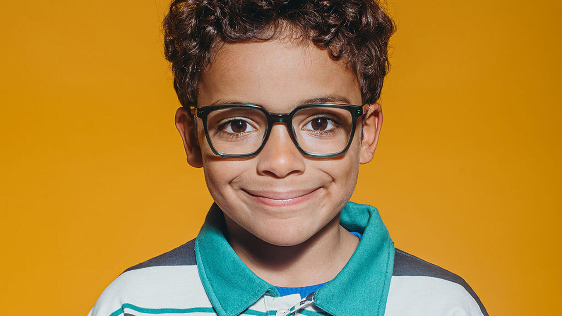 Ein kleiner Junge mit lockigem Haar und großer schwarzer Brille lächelt in die Kamera. Er trägt ein Hemd mit Kragen und blaugrünen und weißen Streifen. Der Hintergrund ist leuchtend gelb.