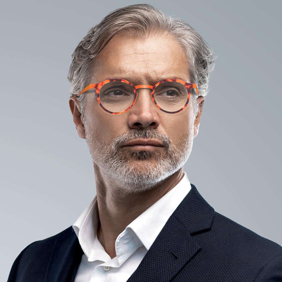 Ein selbstbewusster Mann mittleren Alters mit grauem Haar und ordentlich gestutztem Bart, einer gemusterten orangefarbenen Brille, einem weißen Hemd und einem dunklen Blazer, der vor einem schlichten grauen Hintergrund zur Seite blickt.