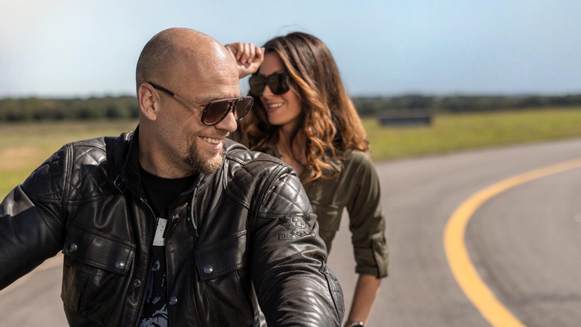 A man and woman wearing sunglasses and leather jackets smile while riding a motorcycle on a curved road with a grassy landscape in the background.