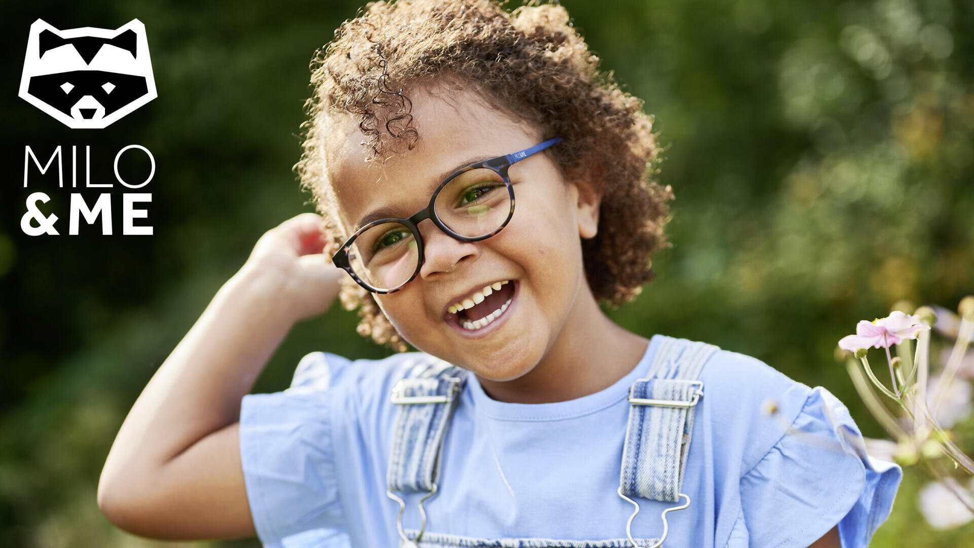 Ein lächelndes Kind mit lockigem Haar und Brille steht in einem blauen Hemd und einem Jeans-Overall im Freien. Das Milo & Me-Logo mit einem Waschbärsymbol befindet sich in der oberen linken Ecke. Im Hintergrund sind helles Grün und Blumen zu sehen.