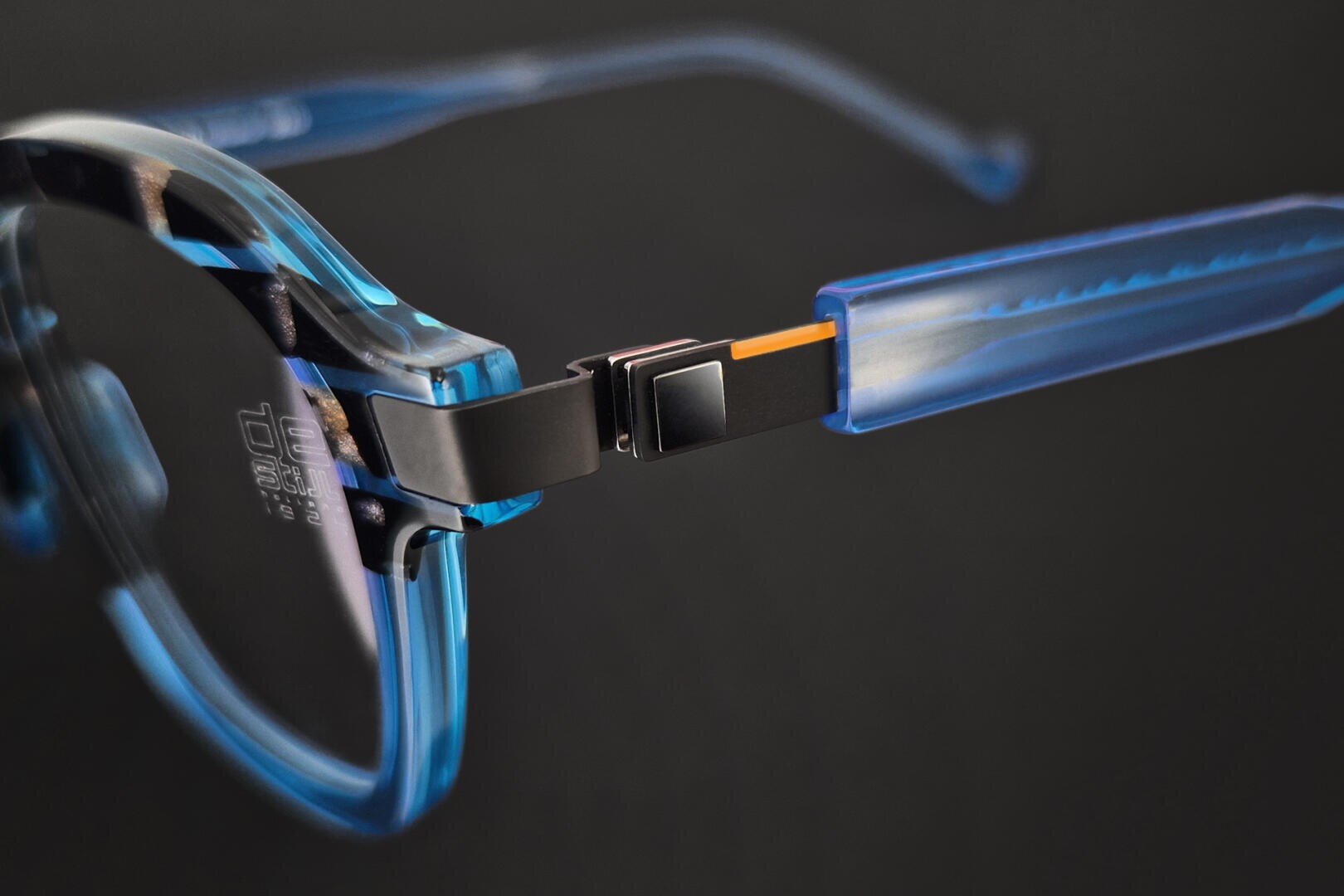 Nahaufnahme einer blauen, durchsichtigen Brille, wobei das Scharnier, an dem der Rahmen auf den Bügel trifft, hervorgehoben wird und Metall- und Kunststoffteile vor einem dunklen Hintergrund zu sehen sind.