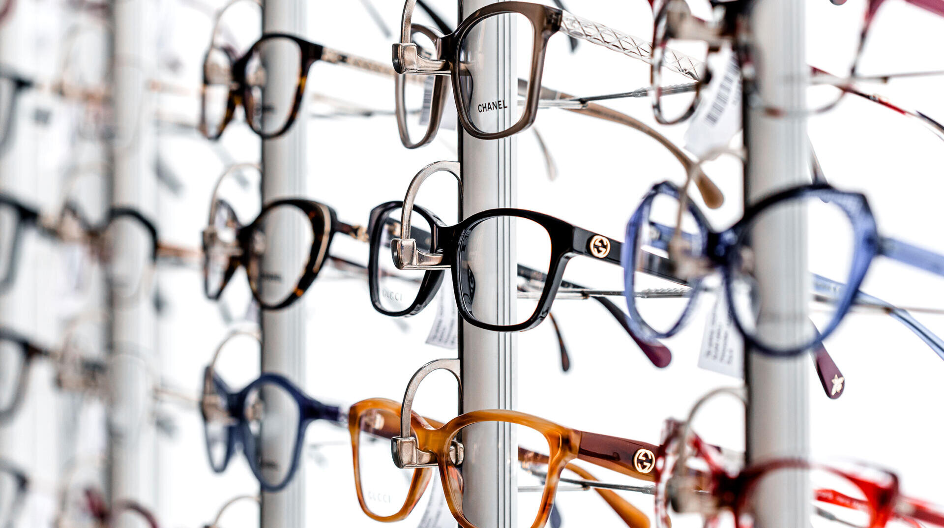 A display rack filled with various eyeglasses in different shapes, sizes, and colors, including brands like Chanel and Gucci, all arranged in rows against a white background.