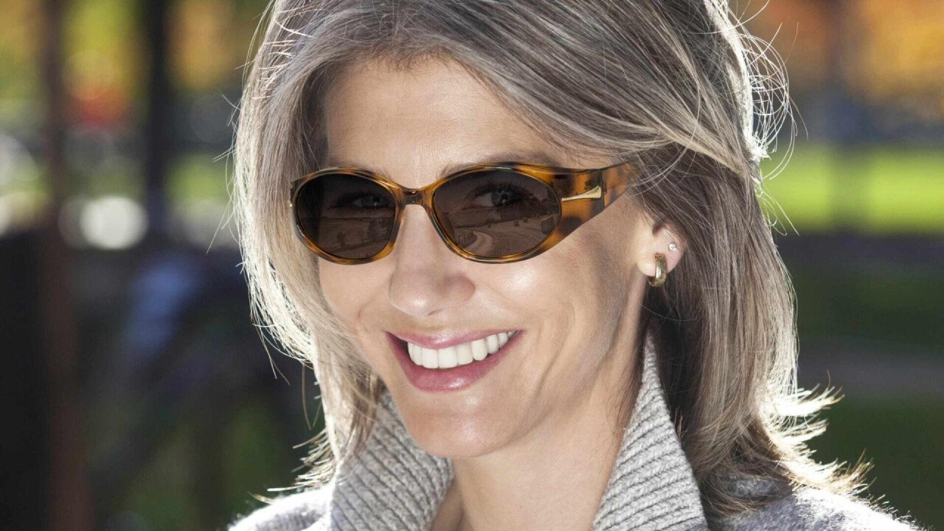 A smiling woman with gray hair wearing sunglasses and a light sweater, standing outdoors with green grass and trees in the blurred background.