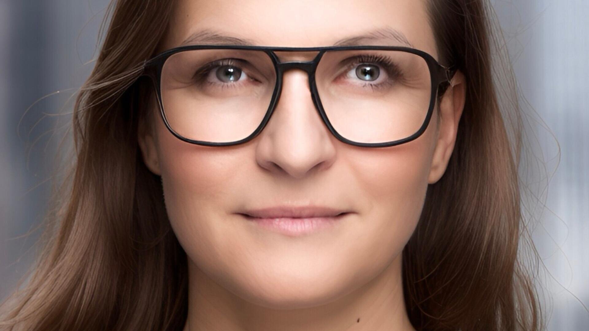 A woman with long brown hair and large, black-rimmed glasses is smiling softly while looking ahead. She is standing in front of a blurred, neutral background.