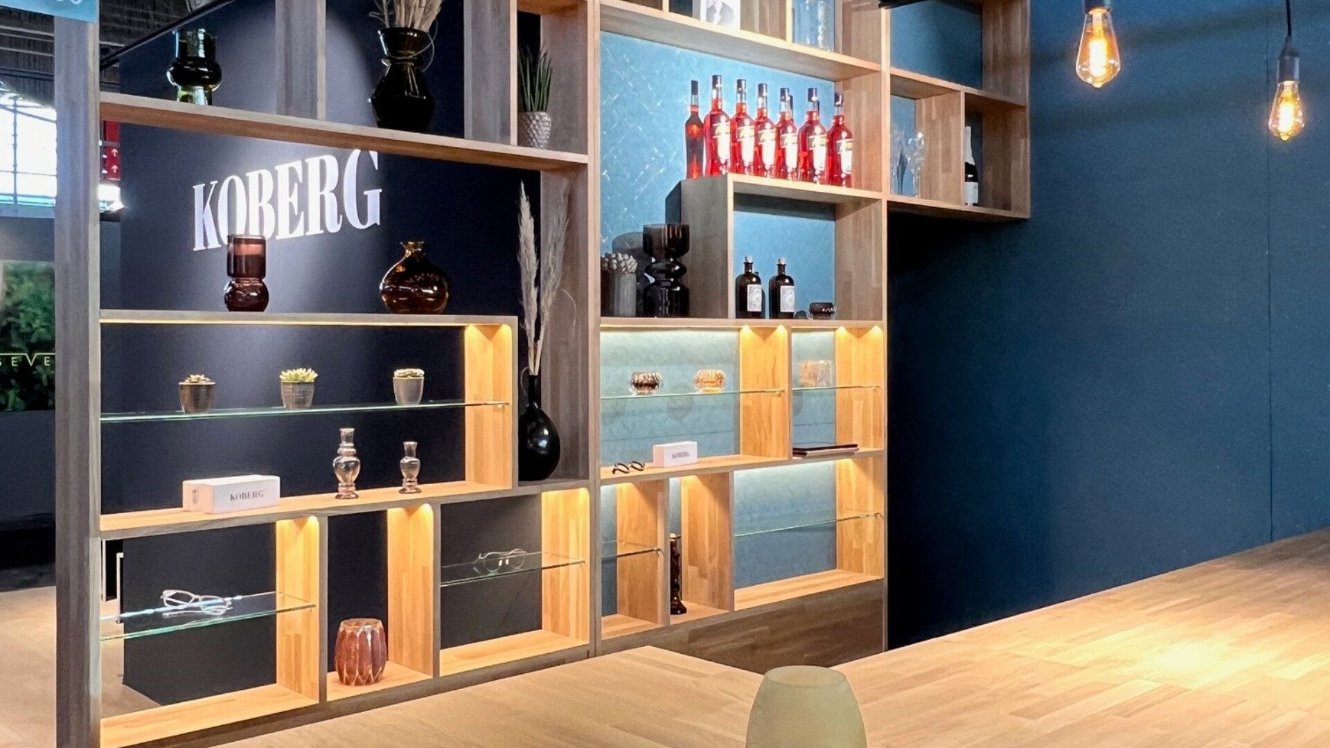 Ein modernes Holzregal mit Glasfächern präsentiert Flaschen, Vasen und Dekoartikel. Warmes Licht hebt die Regale hervor, im Hintergrund ist an einer dunklen Wand ein „KOBERG“-Schild zu sehen.