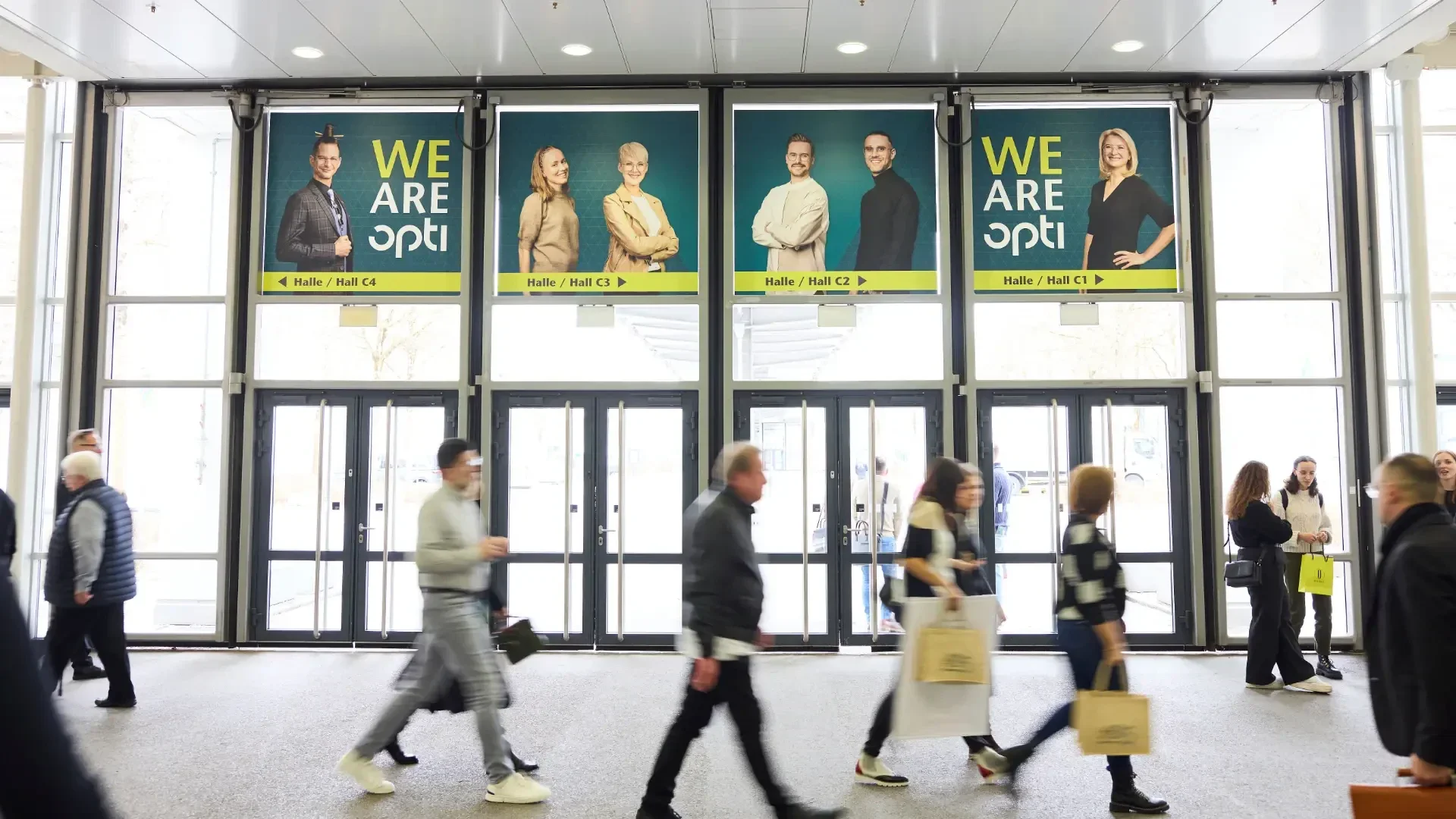 People walk through a bright hallway with large posters above glass doors. The posters feature professionally dressed individuals and the text WE ARE opti along with directions to different halls.