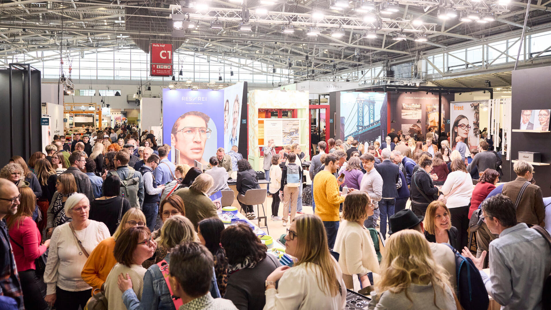 Eine große Menschenmenge versammelt sich in einer belebten Messe- oder Ausstellungshalle voller Stände, Banner und Displays, die alle hell erleuchtet unter einer hohen Decke mit sichtbaren Metallträgern stehen.