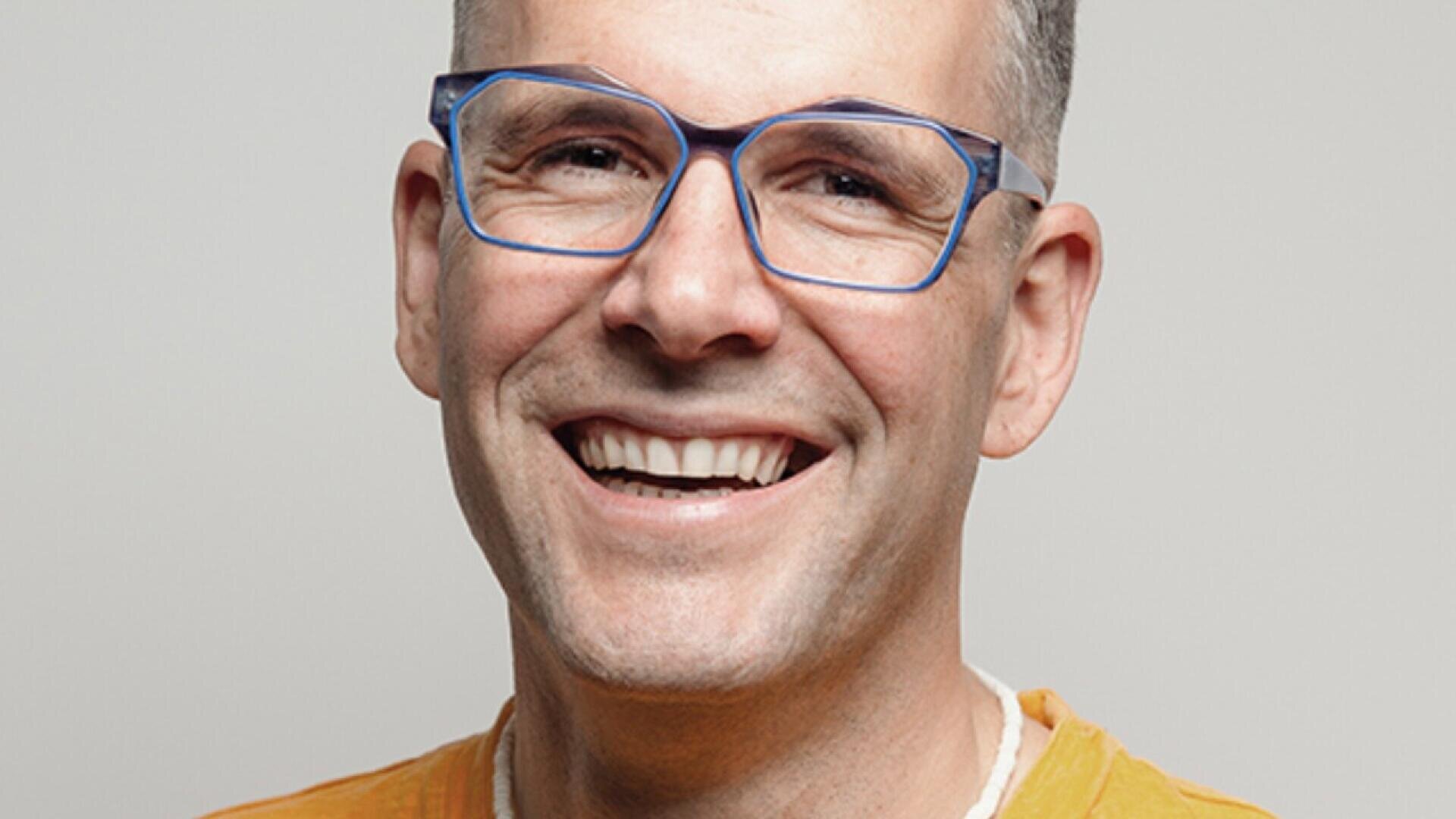 A smiling adult with short gray hair and blue-rimmed glasses wearing an orange shirt, posing against a light gray background.