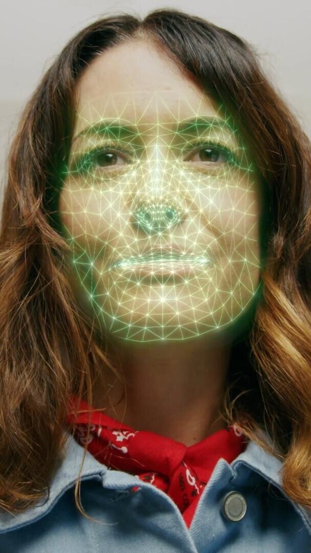 A woman with long brown hair is facing the camera. A green facial recognition grid pattern is overlayed on her face, highlighting her facial features. She is wearing a red scarf and a denim jacket.