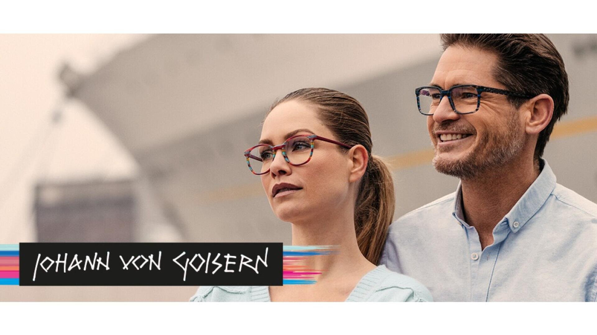 A woman and a man wearing stylish eyeglasses stand outdoors, smiling. The background is blurred, showing a large ship. At the bottom left, Johann von Goisern is written in bold text with colorful accents.