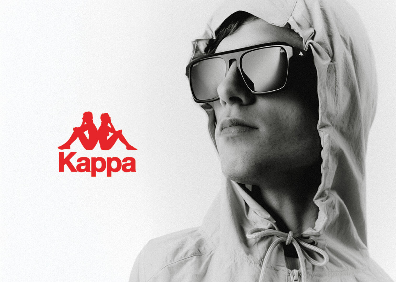 Eine Person, die eine große Sonnenbrille und eine Kapuzenjacke trägt, blickt nach oben. Das rote Kappa-Logo mit zwei Rücken an Rücken sitzenden Figuren und dem Wort "Kappa" erscheint auf der linken Seite des Bildes.