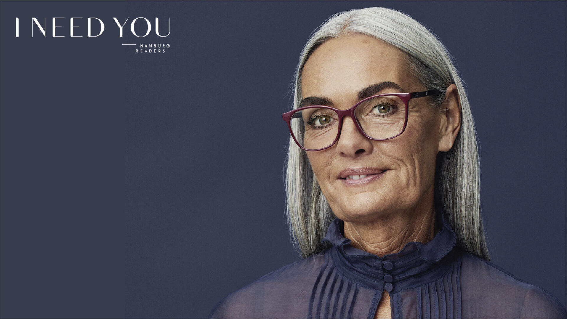 Eine Frau mit langen grauen Haaren, die eine rote Brille und eine durchsichtige dunkle Bluse trägt, lächelt sanft vor einem dunklen Hintergrund. Der Text I NEED YOU HAMBURG READERS erscheint in der oberen linken Ecke.